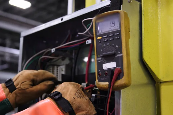 Gloved hands testing wiring in an open electrical enclosure with a digital multimeter