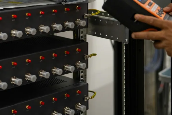 A hand holding a handheld tester beside rack-mounted equipment